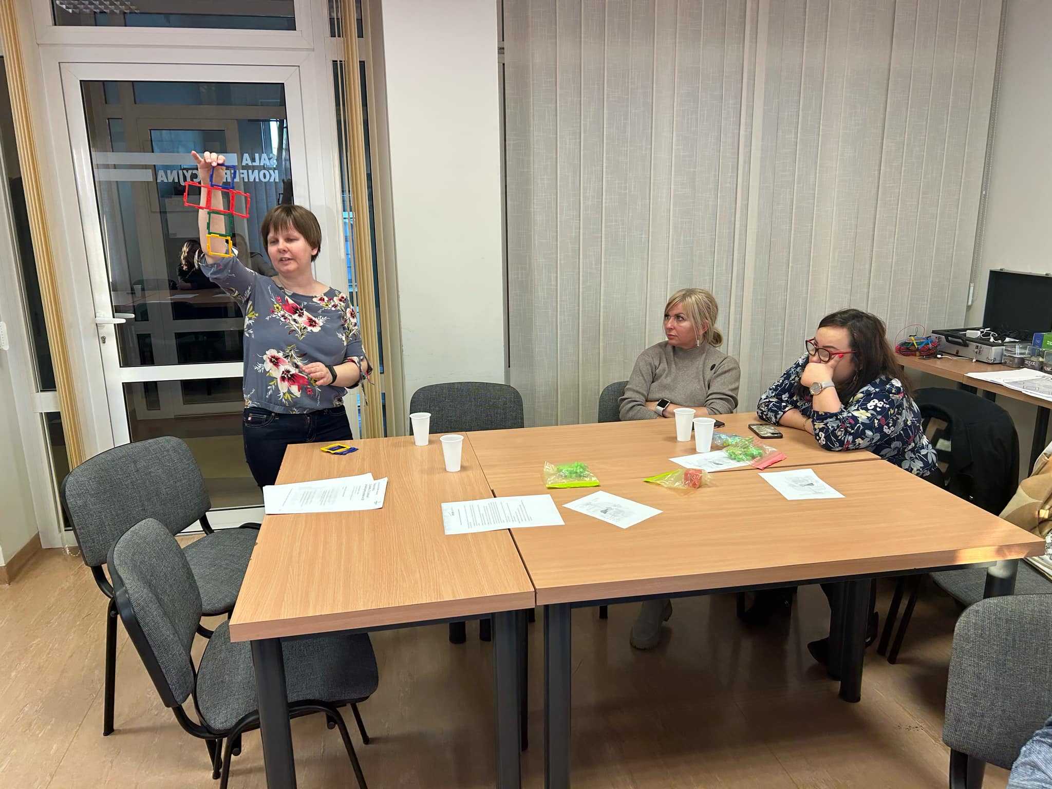 Zdjęcie przedstawia osobę prowadzącą warsztaty. Dwie uczestniczki warsztatów siedzą przy stole na którym, leżą kartki