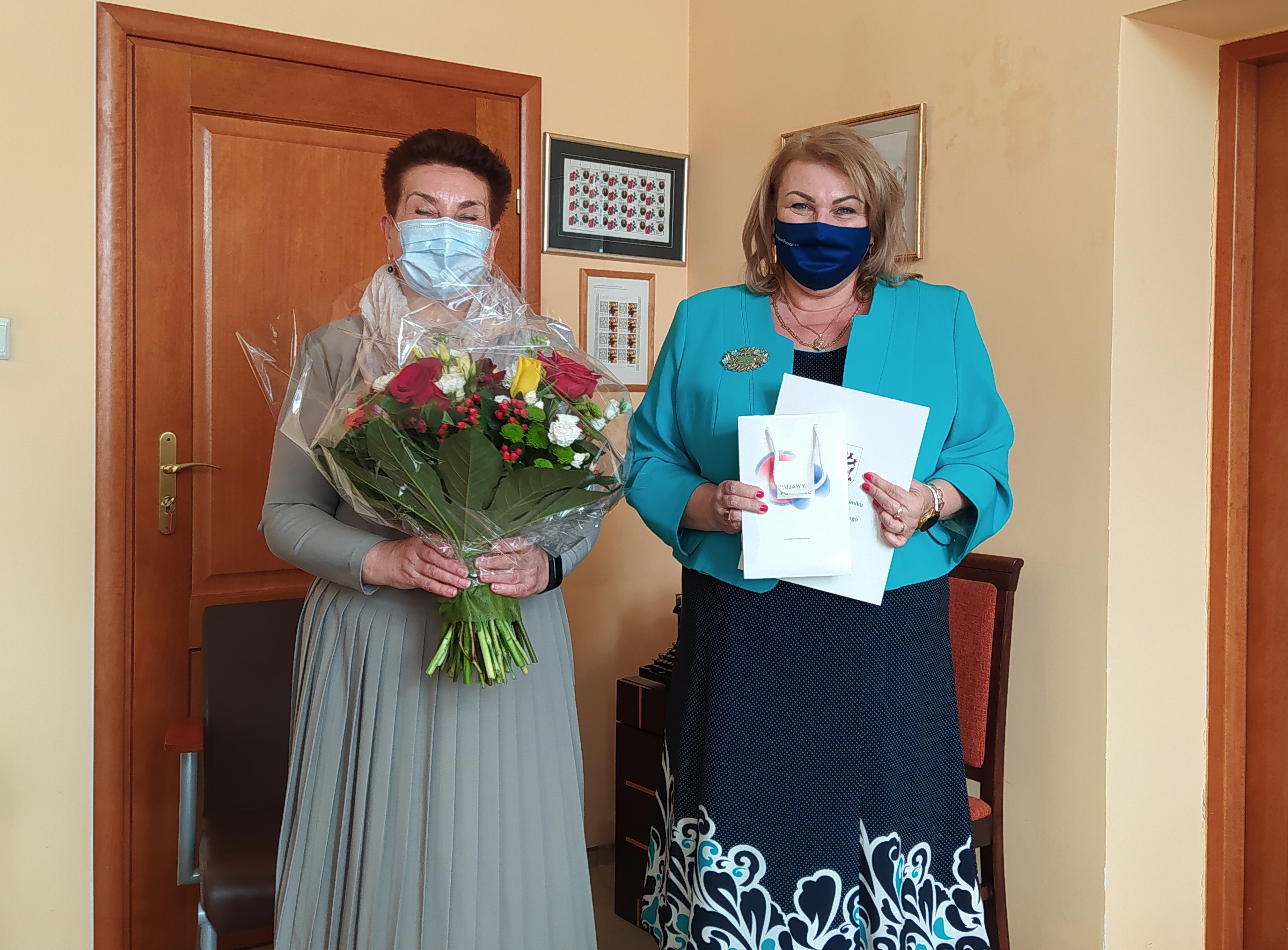 Dwie elegancko ubrane kobiety z maseczkami na twarzach. Jedna trzyma w dłoniach bukiet kwiatów a druga białą teczkę.