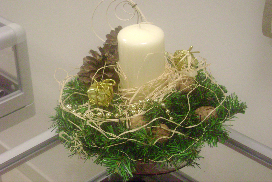 Stroik świąteczny, w którego skład wchodzi: biała świeca, gałązki drzewa iglastego, orzeczy włoskie, szyszki, błyszczące koraliki, miniaturowe prezenty zawinięte w złoty papier i wysuszone trawy.