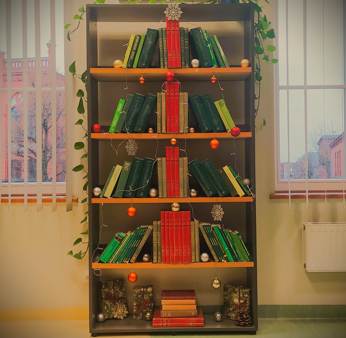 Regał z książkami. Na środku półek ksiązki z czerwonymi grzbietami, tworzące pień choinki. Po bokach po skosie oparte książki z zielonymi grzbietami, stanowiące gałęzie choinki.