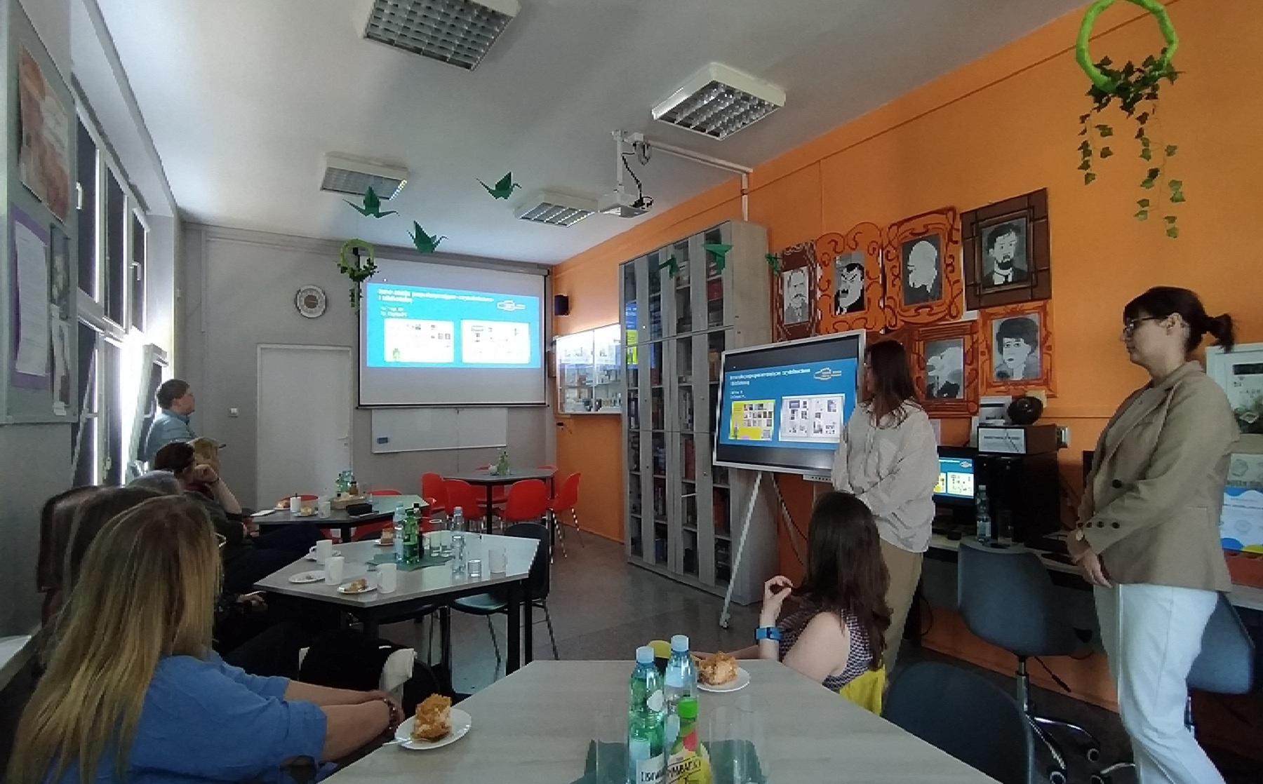 Sala biblioteki szkolnej, na wprost ekran z wyświetlona prezentacją, ludzie siedzący przy stolikachj i stojący po prawej stronie.