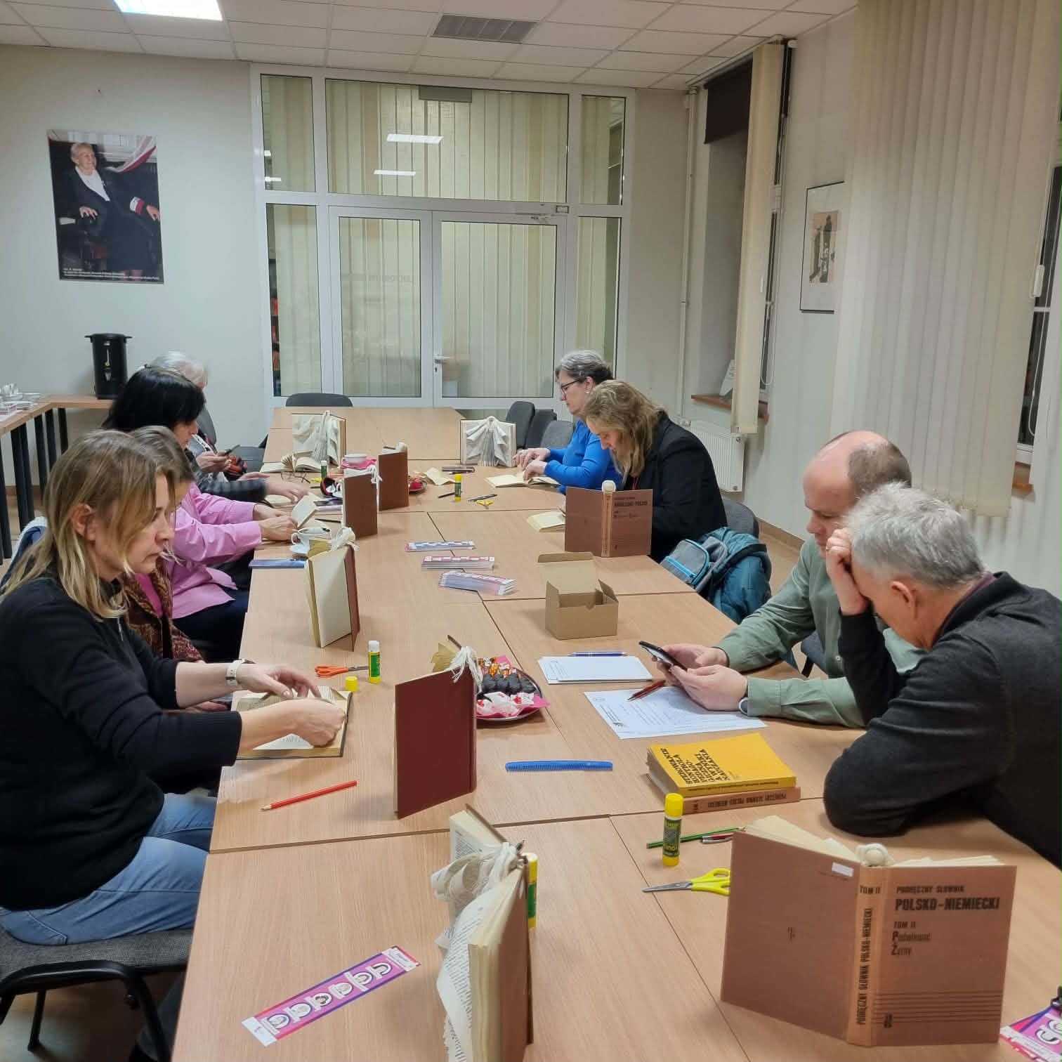 Grupa ludzi siedzi przy stołach w sali konferencyjnej Biblioteki Pedagogicznej w Toruniu. Przed nimi stoją stare książki, uczestnicy spotkania wykonują prace plastyczne.