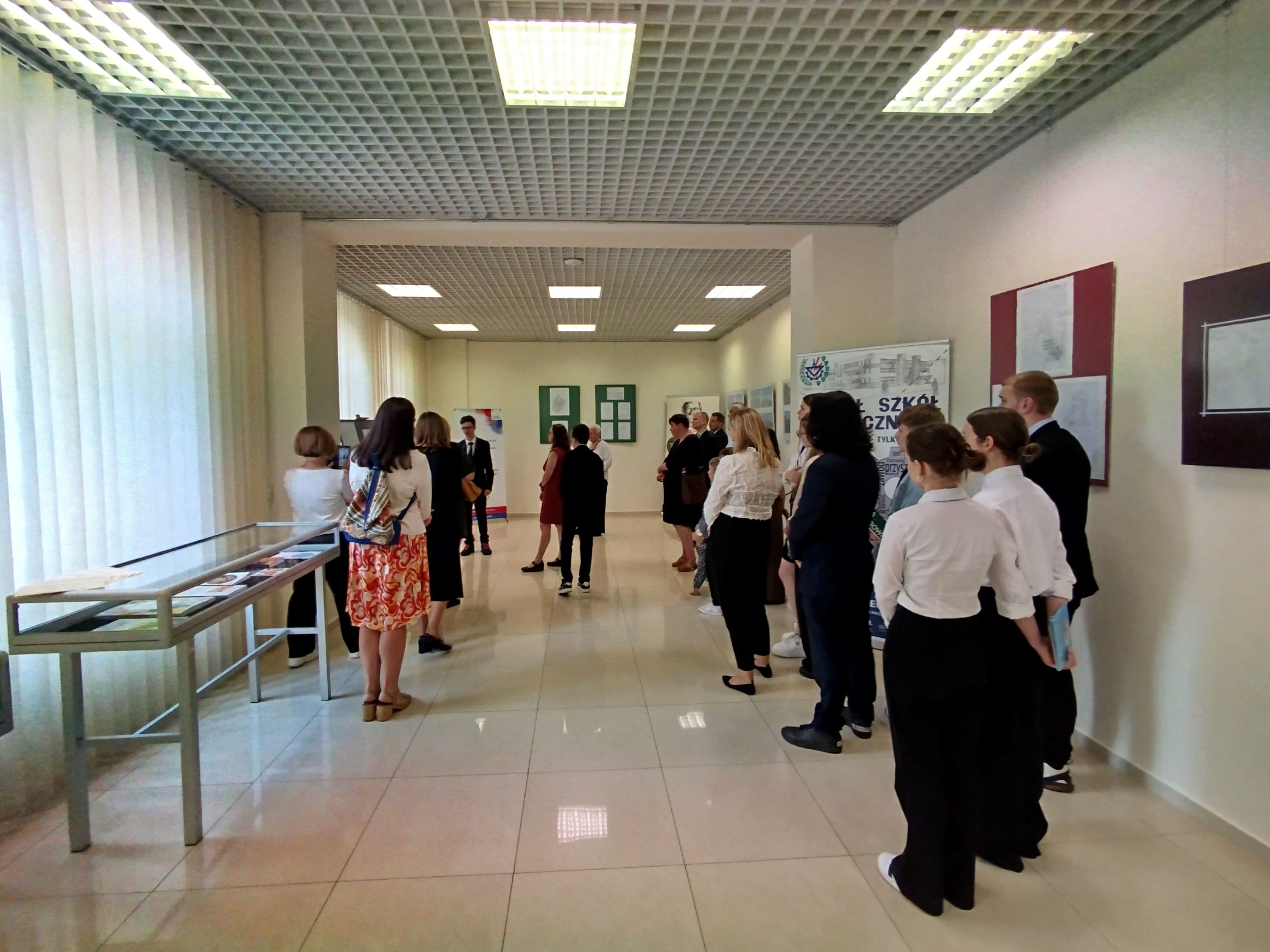 Grupa ludzi stoi w sali wystawowej Biblioteki Pedagogicznej. Na ścianach wiszą prace plastyczne. Po lewej stronie stoją gabloty.