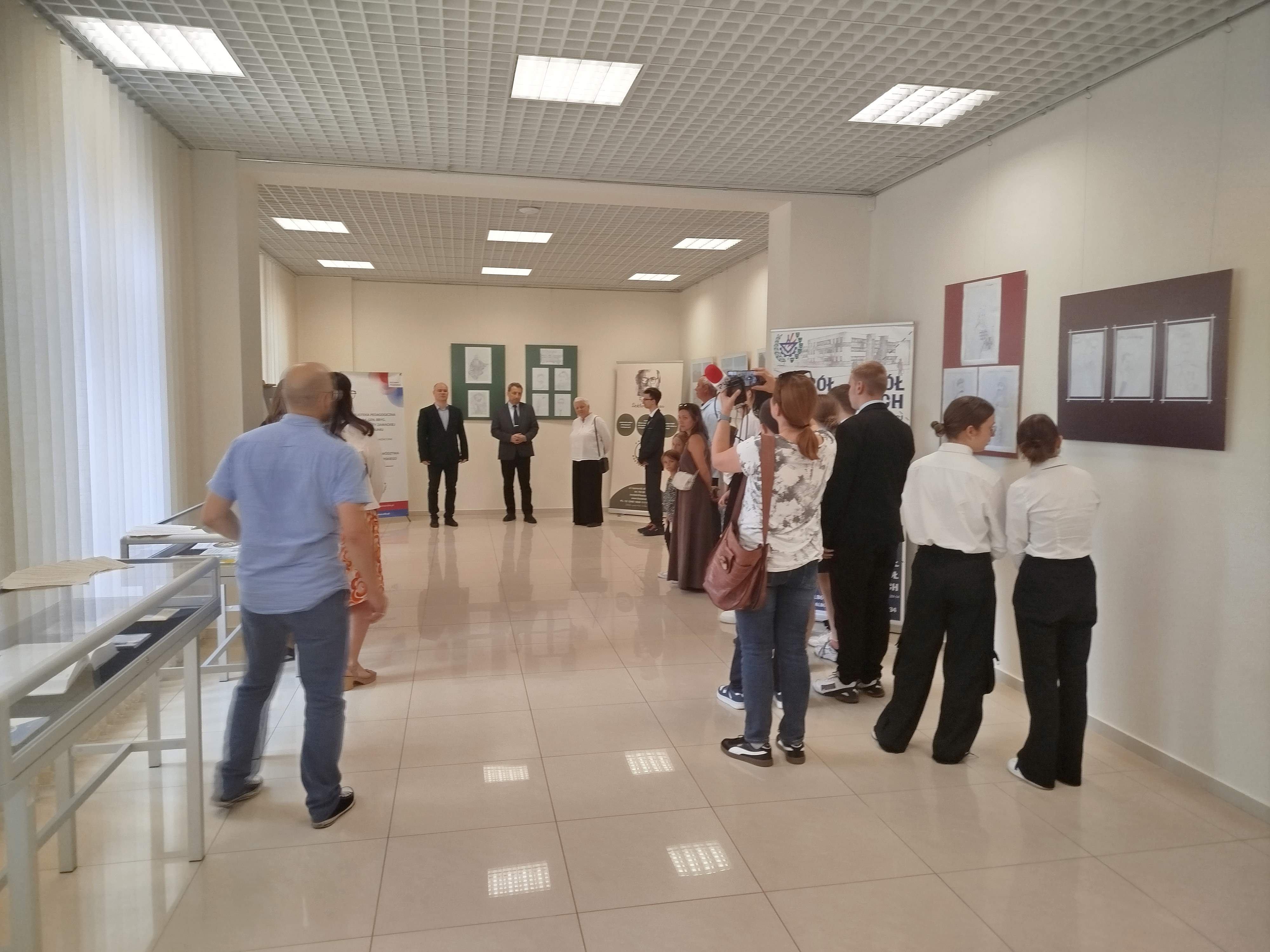 Grupa ludzi stoi w sali wystawowej Biblioteki Pedagogicznej. Na ścianach wiszą prace plastyczne. Pod ścianą na przeciw wejścia stoi trzech mężczyzn, kobieta i nastolatek.