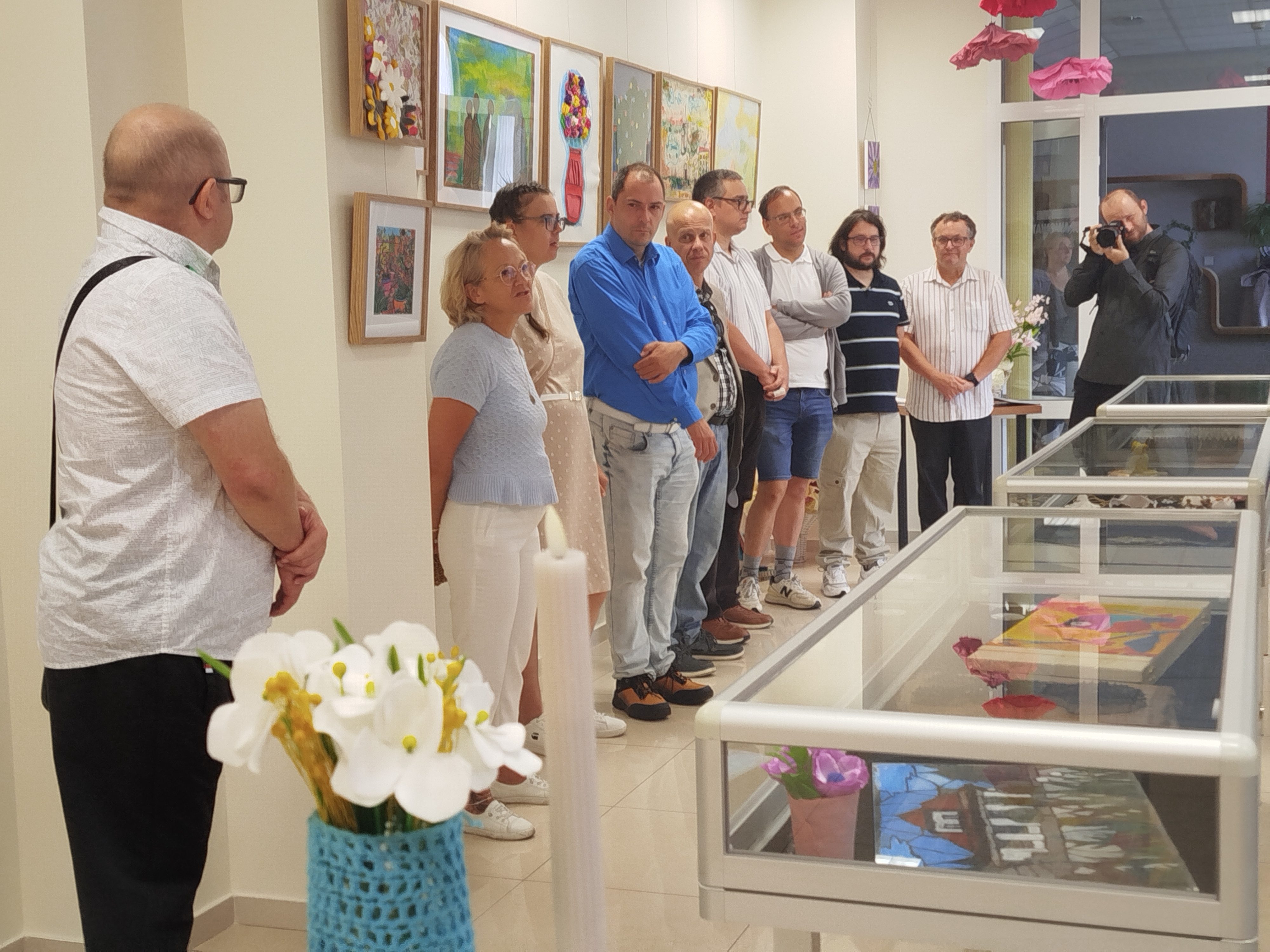 Grupa ludzi w&nbsp;sali wystawowej, na ścianach wiszą kolorowe prace plastyczne, po srodku sali stoją gabloty wystawowe.