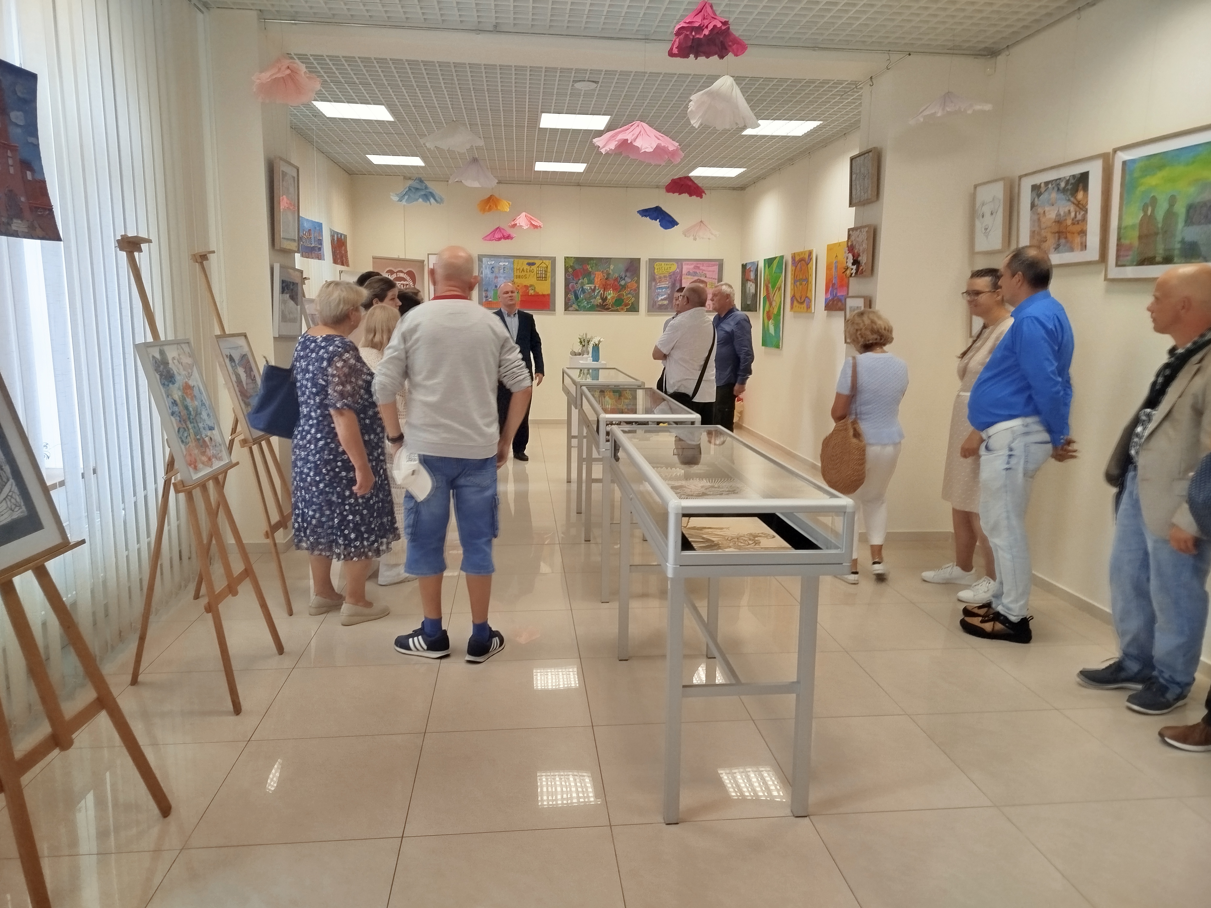 Grupa ludzi w&nbsp;sali wystawowej, na ścianach wiszą kolorowe prace plastyczne, po srodku sali stoją gabloty wystawowe.